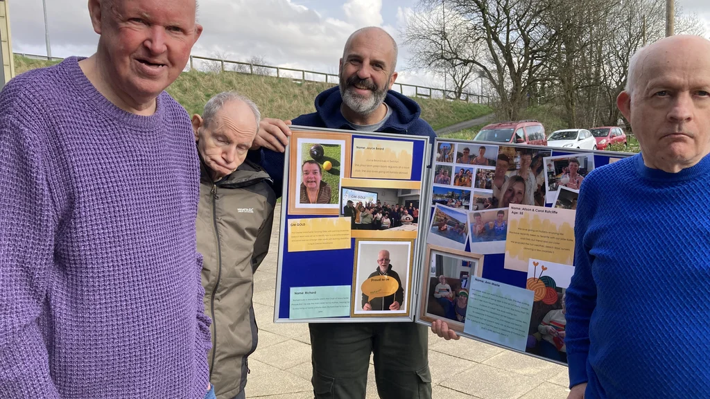 Four men hold up a noticeboard with pictures and exhibition information