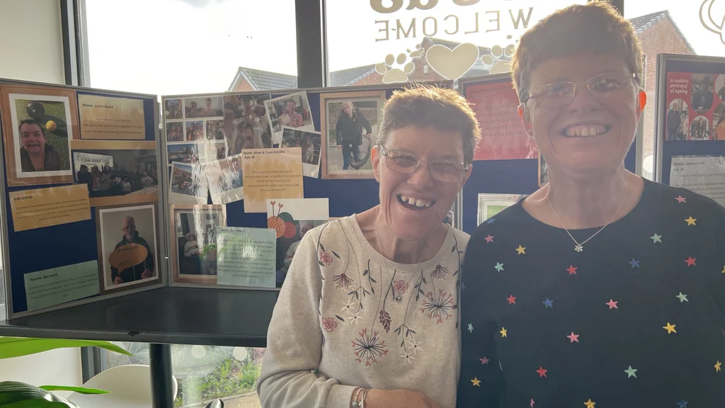 Two women smiling in front of an exhibition noticeboard 